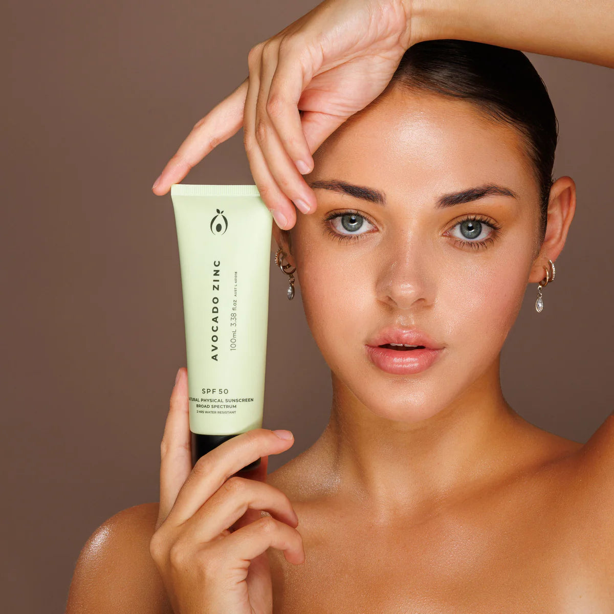 Woman holding a tube of Avocado Zinc sunscreen against a brown background. Avocado Zinc SPF 50 Natural Physical Sunscreen 100ml