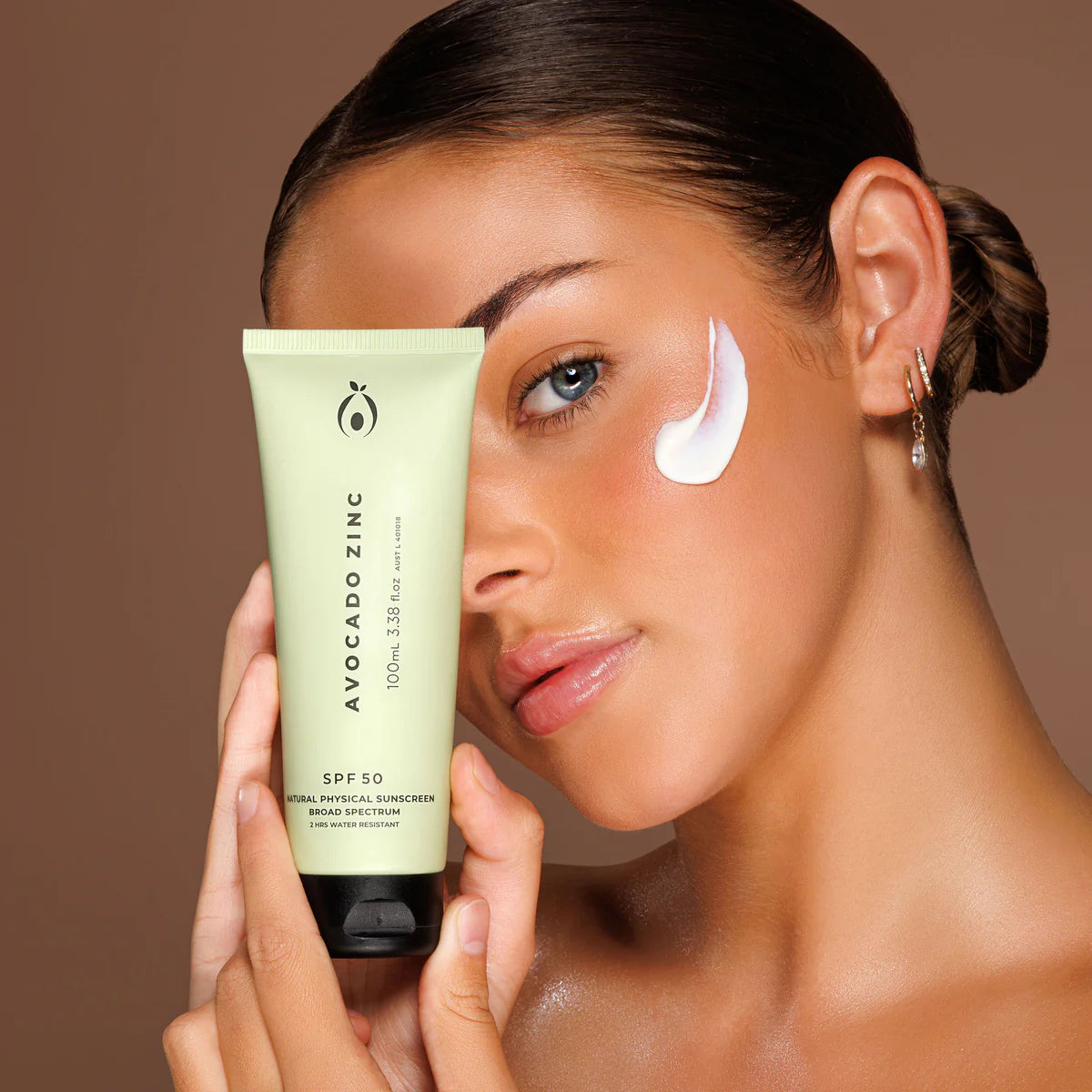 Woman applying sunscreen with a tube labeled 'Avocado Zinc' on a brown background. Avocado Zinc SPF 50 Natural Physical Sunscreen 100ml