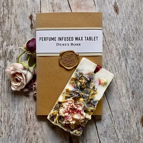 A perfume-infused wax tablet labeled 'Dusty Rose' placed on top of its kraft paper packaging, styled on a rustic wooden surface with dried rose petals and a pale rose bud.