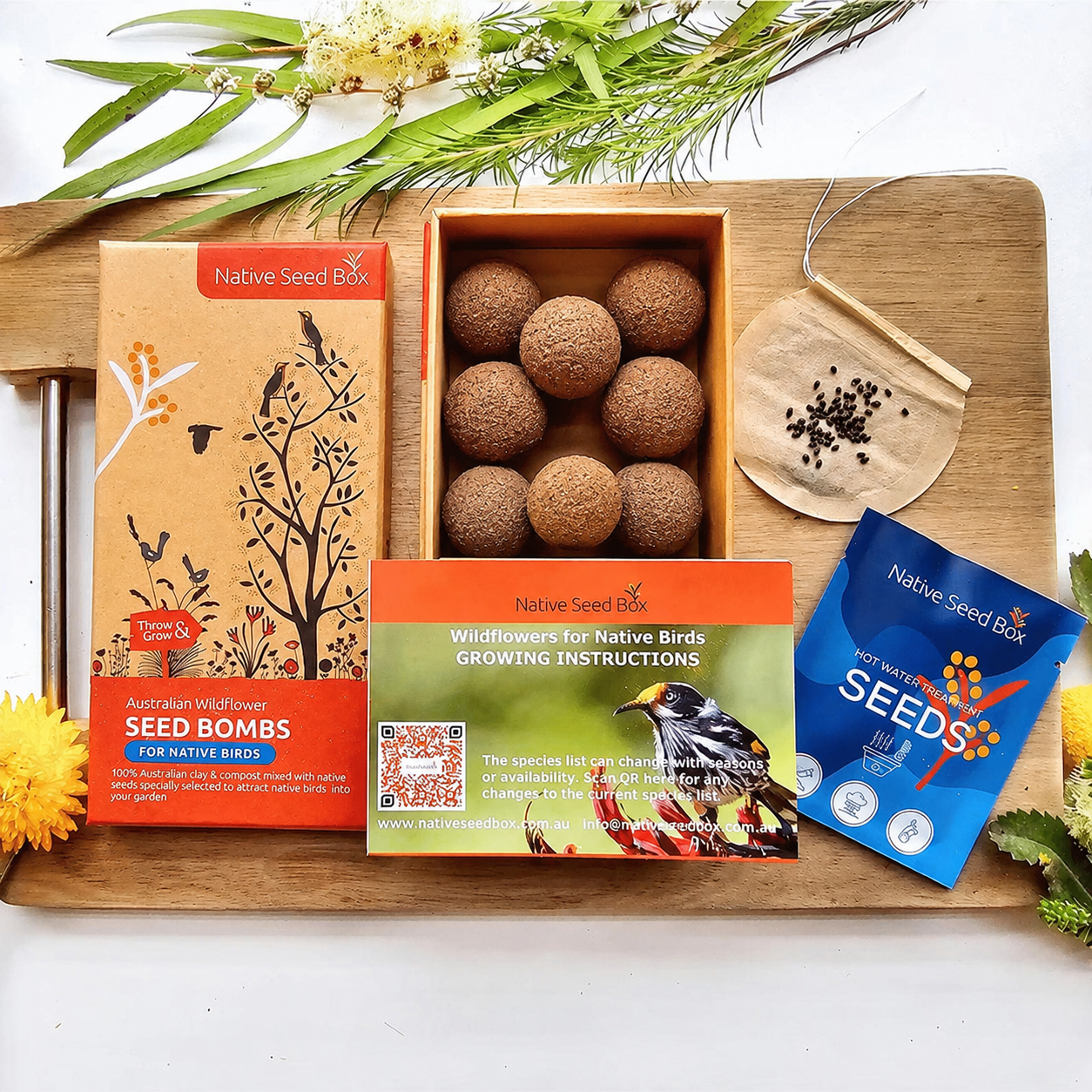 Native Seed Box bird seed kit arranged on a wooden board with greenery and flowers. The set includes a seed bombs box with round brown seed bombs, a seed packet, a growing instructions card, and a small seed scoop.