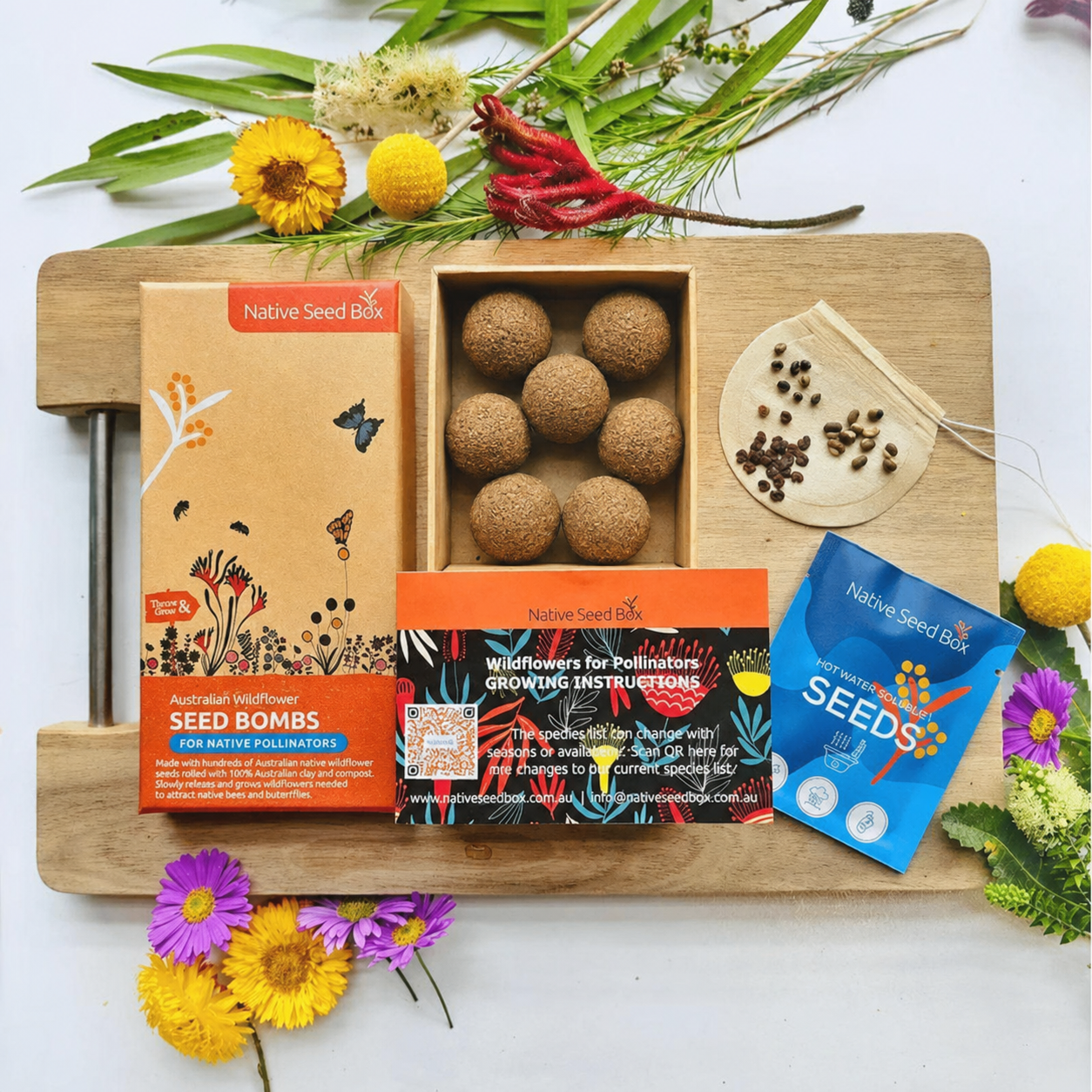 A styled flat-lay of a Native Seed Box Seed Bombs kit on a wooden board, featuring a kraft paper box, an open tray of round brown seed bombs, an orange growing instructions card, a blue seeds packet, and decorative wildflowers and greenery on a white background.
